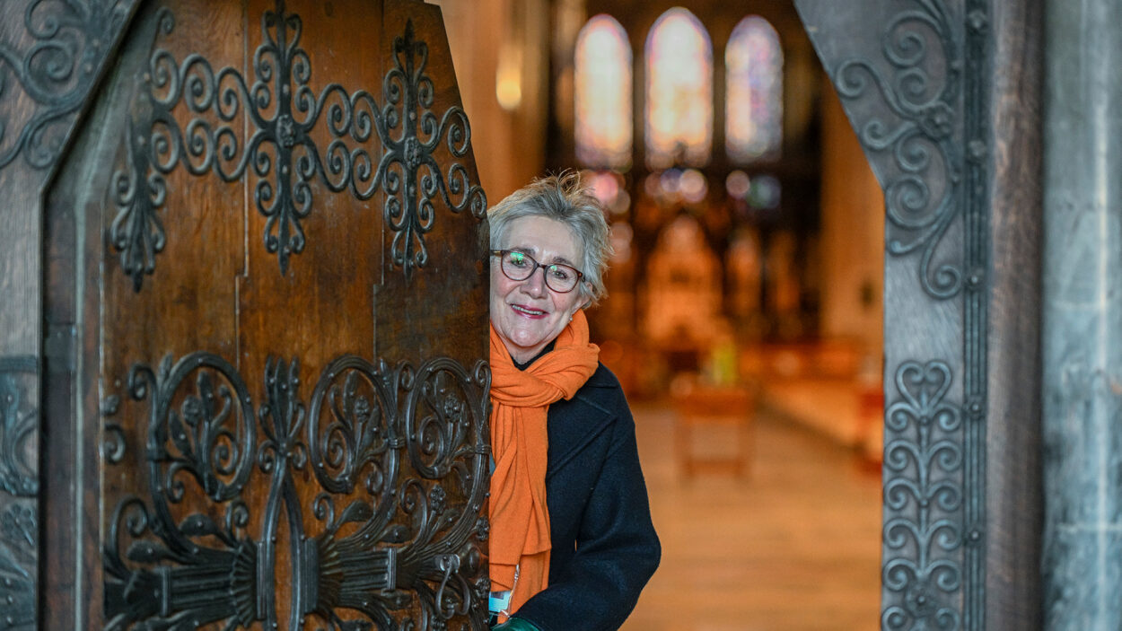 A friendly looking woman opening am ornate carved door revealing a warm cathedral interior